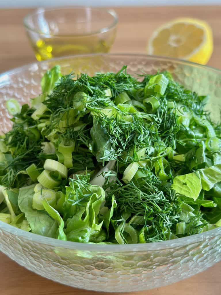 Greek Green Salad Maroulosala in a glass bowl with lemon and olive oil behind it