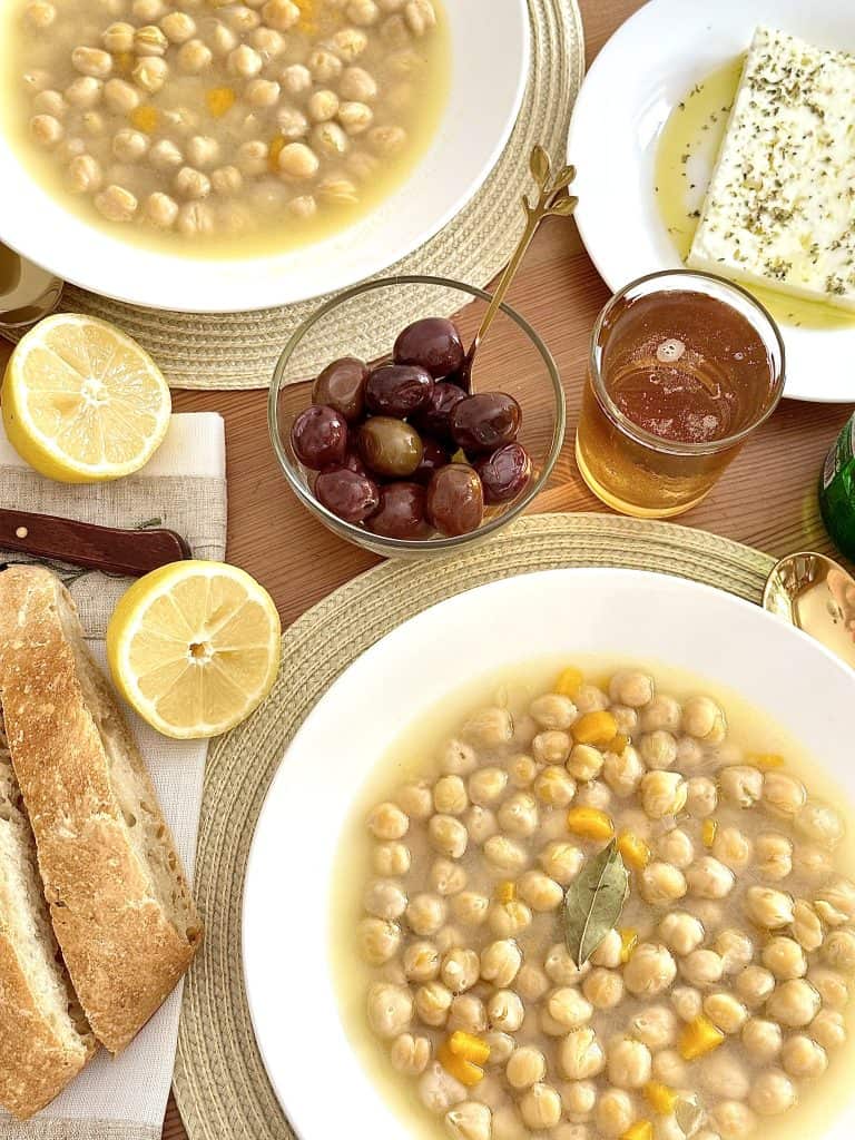 Two bowls of Instant Pot Greek Chickpea Soup served on a wooden table with lemon, bread, olives, and a glass of beer on the side