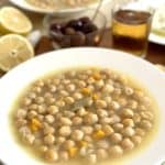 Instant Pot Greek Chickpea Soup Revithia served in a white bowl on a wooden table with lemons and olives, and a glass of beer on the side