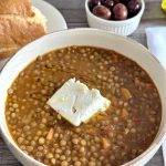 The best Greek lentil soup recipe ever (Fakes Soupa) in a bowl with bread, olives, vinegar, and olive oil next to it